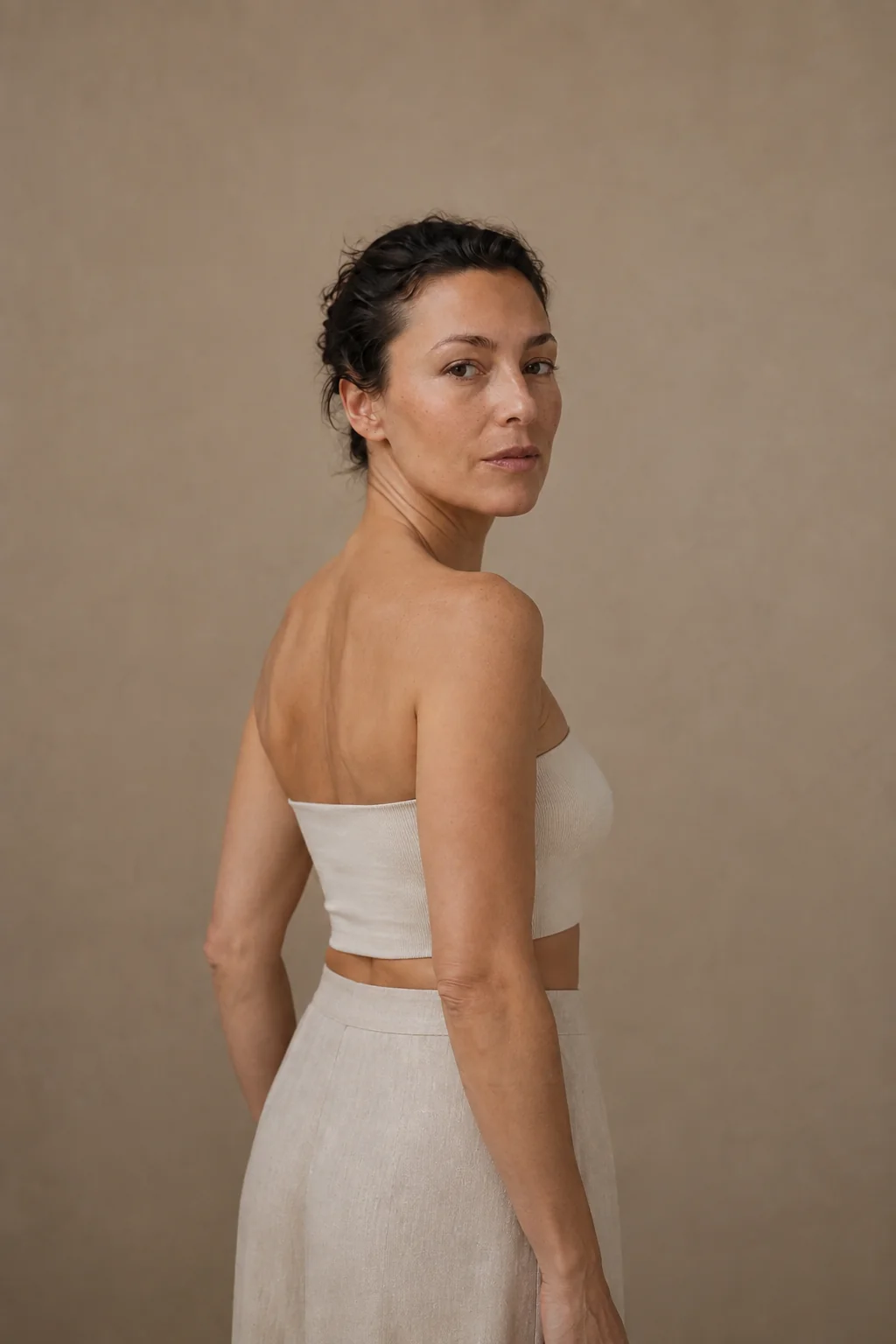 AI-generated editorial portrait of a woman looking over her bare shoulder, wearing an ivory bandeau and linen skirt against a warm taupe backdrop.