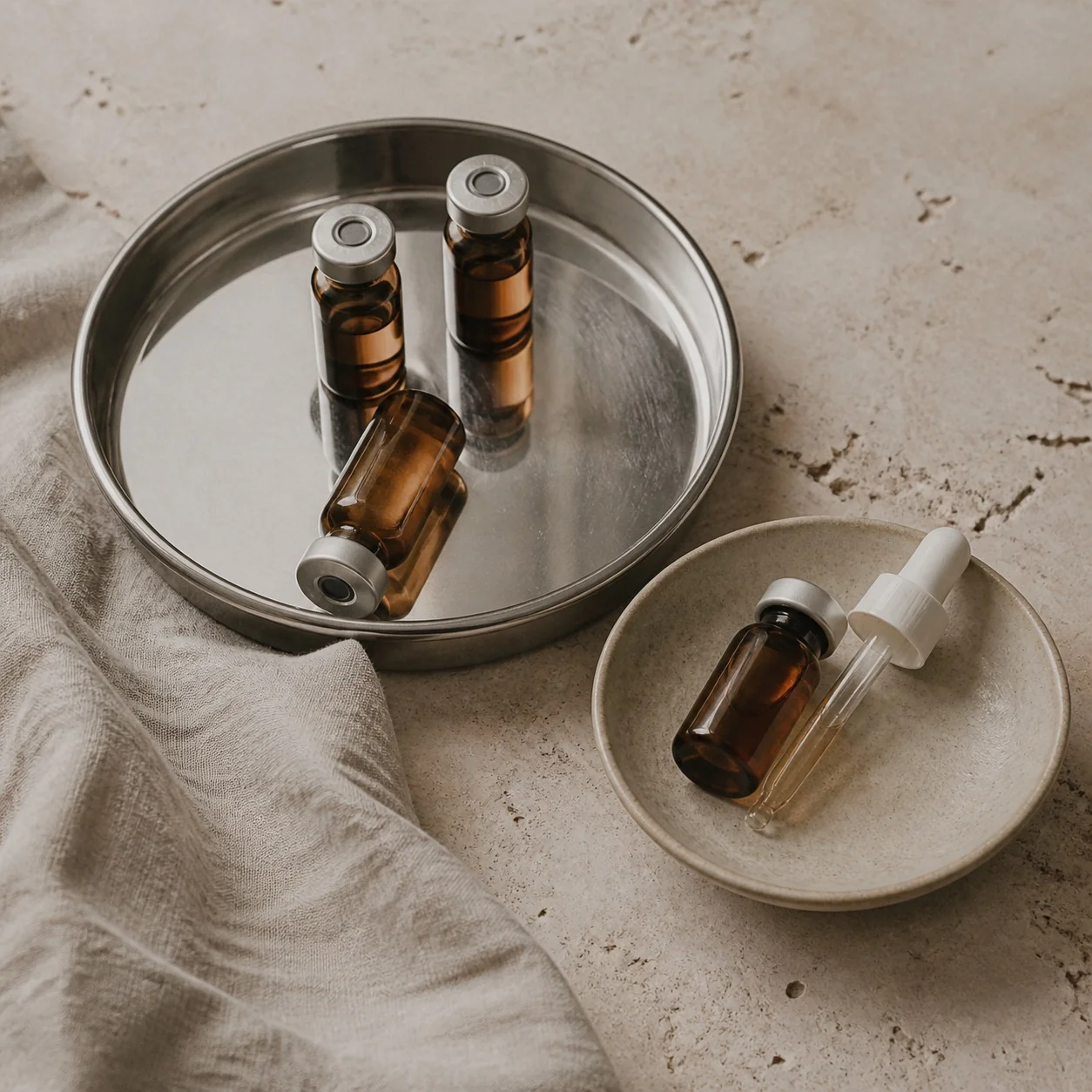 Overhead still life of amber vials, a steel tray and a ceramic dish on pale stone.